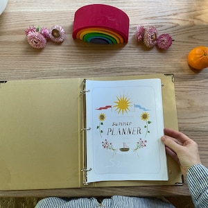 May include: A gold binder with a white page open to a summer planner with a sun, sunflowers, and a sailboat. The text on the page reads "Summer Planner".