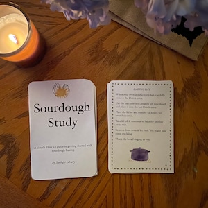 May include: A white card with the title "Sourdough Study" and a simple guide to sourdough baking. The card is on a wooden table with a lit candle and a bouquet of purple flowers.