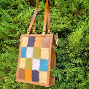 Può includere: Una borsa tote in pelle marrone con un design patchwork sul davanti. Il patchwork presenta quadrati di vari colori, tra cui blu, verde, arancione e beige. La borsa ha cinghie in pelle marrone e una chiusura con cerniera.