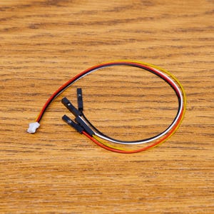 Könnte beinhalten: Ein Satz mehrfarbiger Elektrokabel mit schwarzen Steckern und einem weißen Stecker. Die Kabel sind rot, gelb, schwarz und weiß. Die Kabel sind in einer gebogenen Form auf einer Holzoberfläche angeordnet.
