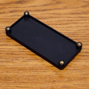 Peut inclure: Un plateau rectangulaire noir avec des coins arrondis et quatre vis dorées. Le plateau est posé sur une surface en bois avec un motif de grain de bois naturel. Le plateau est de conception simple et moderne.