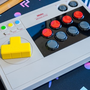 Könnte beinhalten: Ein weißer und grauer Arcade-Stick mit einem gelben Joystick und roten und grauen Knöpfen. Der Arcade-Stick hat den Text "Smitho ARCADE STICK" auf der Vorderseite.