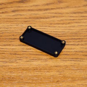 May include: A rectangular black object with rounded corners and four gold-colored screws. The object is set against a wooden background with a natural grain pattern. The item appears to be a small electronic component or housing.