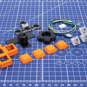Könnte beinhalten: Sortiment an orangefarbenen und schwarzen mechanischen Tastaturkappen und -schaltern, zusammen mit einem schwarzen Knopf und einem Satz Drähte. Die Artikel sind auf einer blauen, gerasterten Oberfläche angeordnet.