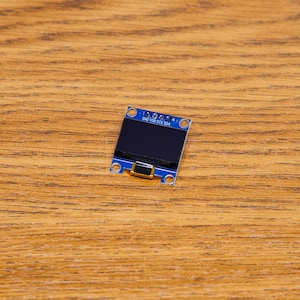 May include: A small, rectangular blue OLED display module with a black screen and gold-coloured accents. The module has four mounting holes and is resting on a wooden surface. The module has text printed on it.