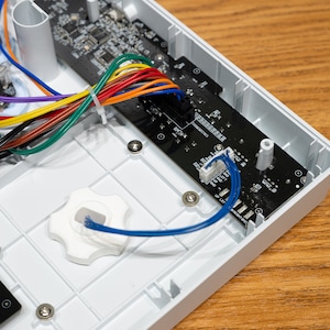 Könnte beinhalten: Nahaufnahme des Inneren eines weißen elektronischen Geräts mit einer Platine, Drähten und einem weißen Knopf. Die Drähte sind farbcodiert mit Rot, Blau, Grün, Gelb und Schwarz.