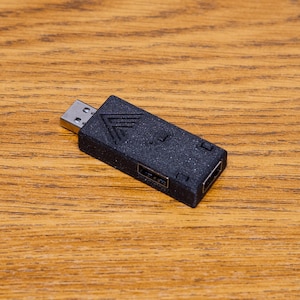 Puede incluir: Un adaptador USB negro con un conector USB plateado y dos puertos. El adaptador tiene una superficie texturizada y un diseño geométrico en la parte delantera. Está sobre una superficie de madera.