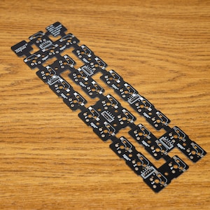 May include: A collection of black circuit boards with white markings and text, arranged diagonally on a wooden surface. The boards have rectangular and rounded sections with various holes and components. The background is a light brown wood grain.