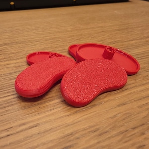 May include: Four small, kidney-shaped red plastic components with a textured top surface. Two of the pieces are positioned face-up, showing a smooth underside with a central circular mounting post, while the other two show the textured top.
