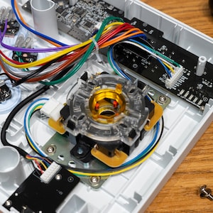 Peut inclure: Gros plan de l'intérieur d'un boîtier d'arcade blanc avec un joystick, des boutons et du câblage. Le joystick est en plastique transparent avec un centre doré et est connecté à une carte de circuit imprimé noire.