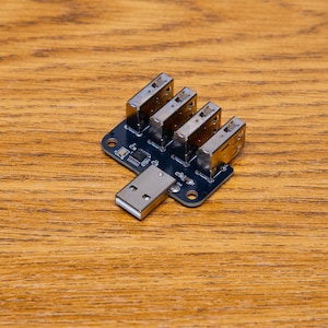Könnte beinhalten: Eine kleine, dunkelblaue Leiterplatte mit vier silbernen USB-Anschlüssen und einem USB-Anschluss. Die Platine liegt auf einer Holzoberfläche.