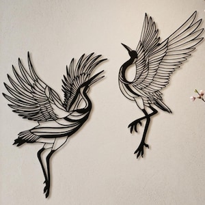 May include: Two black metal crane wall art pieces with outstretched wings are mounted on a light-colored wall. The cranes are in a Japanese-inspired dining room, with a dark wood table set with plates, chopsticks, and a vase of flowers.