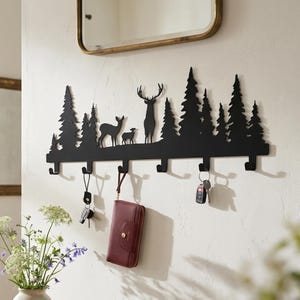 May include: A black metal wall-mounted coat rack featuring a silhouette of a forest with pine trees, a deer, and a fawn. The rack has seven hooks for hanging items, with keys and a maroon wallet currently suspended from the hooks on the wall.