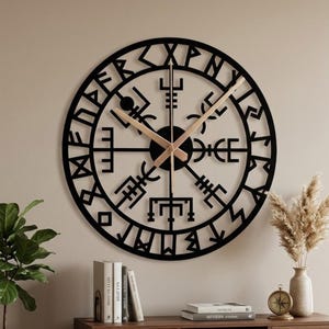 Può includere: Un grande orologio da parete rotondo nero con un design geometrico unico. Le lancette dell'orologio sono marroni chiare. L'orologio è montato su una parete beige sopra un mobile in legno. Il mobile ha libri e oggetti decorativi.