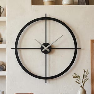 Op de afbeelding: Een grote ronde wandklok met een zwart metalen frame en een kruisdraadontwerp dat voorbij de buitenste ring steekt. De klok heeft een centrale naaf met twee zilverkleurige wijzers die de tijd aangeven tegen een effen, lichte muur.