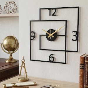 Puede incluir: Un moderno reloj de pared de metal negro con manecillas doradas y un centro circular negro. El reloj presenta un diseño único de cuadrado dentro de un cuadrado con los números 3, 6, 9 y 12. El reloj está montado en una pared blanca.