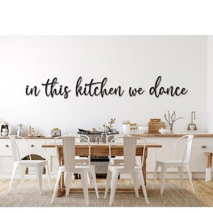 Könnte beinhalten: Küchenszene mit einem Holztisch und weißen Stühlen. Der Schriftzug "in this kitchen we dance" ist in schwarzer Schrift an der weißen Wand angebracht. Die Küche hat weiße Schränke und eine helle Holz-Arbeitsplatte.