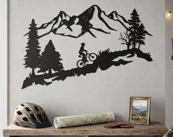 May include: Black metal wall art depicting a mountain range with a cyclist on a mountain bike. The artwork includes silhouetted trees and grass. A bicycle helmet, map, and framed picture sit on a wooden shelf below the art.