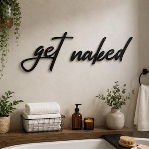 May include: A black wall-mounted sign featuring the cursive text get naked. The lettering is constructed from a solid material and is displayed horizontally against a neutral-coloured wall above a bathtub area with various bathroom accessories.