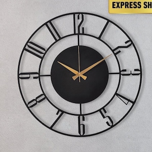 May include: A black metal wall clock with Roman numerals and gold-colored hands. The clock is circular and mounted on a light gray wall above a black leather sofa. The clock's design is modern and minimalist.