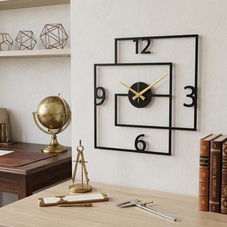 May include: A contemporary black metal wall clock with gold hands and a black circular centre. The clock has a unique square-in-square design with the numbers 3, 6, 9, and 12. The clock is mounted on a white wall.