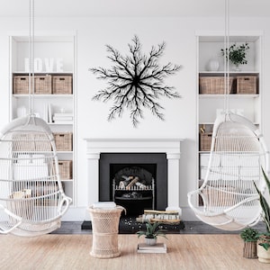 Op de afbeelding: Een zwart, stervormig kunstwerk hangt boven een open haard in een lichte, luchtige woonkamer. Twee witte rotan hangstoelen en een geweven vloerkleed dragen bij aan de natuurlijke esthetiek van de kamer. Planken met manden en planten maken de inrichting compleet.