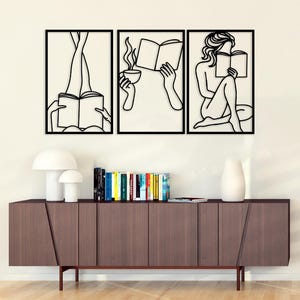Puede incluir: Conjunto de arte mural de tres piezas con dibujos lineales negros de figuras leyendo libros. La obra de arte representa a una persona con las piernas levantadas, otra sosteniendo una taza y una tercera sentada. El conjunto está sobre un mueble de madera.