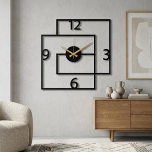 May include: A contemporary black wall clock with a distinctive geometric design. The clock has a black circular face with gold-coloured hands and the numbers 3, 6, 9, and 12. It is mounted on a light grey wall.