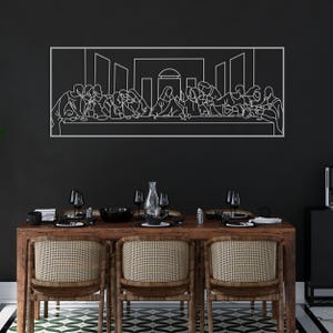 May include: A white metal wall art piece depicting the Last Supper scene, mounted on a black wall. The artwork is above a wooden dining table with woven chairs, set with glassware and tableware. The room has a modern aesthetic.