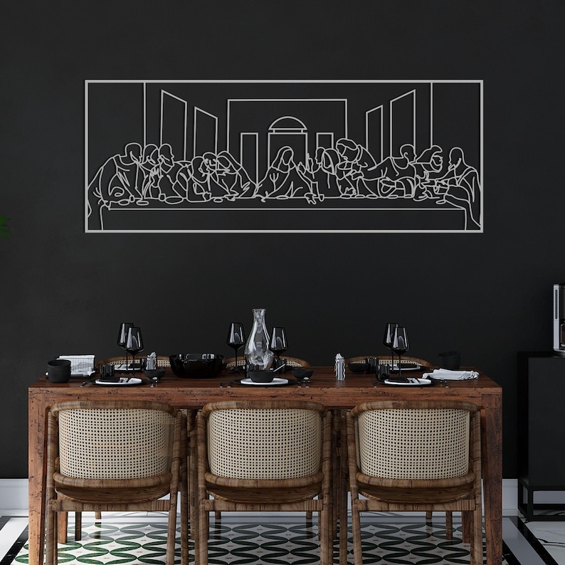 May include: A white metal wall art piece depicting the Last Supper scene, mounted on a black wall. The artwork is above a wooden dining table with woven chairs, set with glassware and tableware. The room has a modern aesthetic.