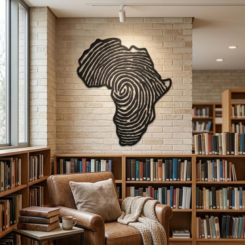 Op de afbeelding: Een zwart-wit kunstwerk in de vorm van het Afrikaanse continent, met een vingerafdrukontwerp. Het kunstwerk is gemonteerd op een bakstenen muur in een bibliotheek, met boekenplanken en een leren fauteuil.