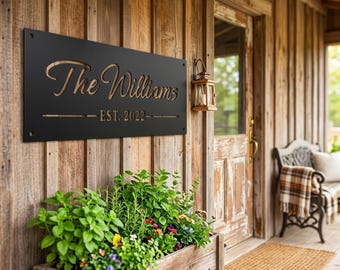 Puede incluir: Un letrero rectangular de metal negro con letras recortadas que dicen The Williams y EST. 2022. El letrero cuenta con cuatro orificios de montaje, uno en cada esquina, y está montado en una pared de madera vertical sobre una jardinera.