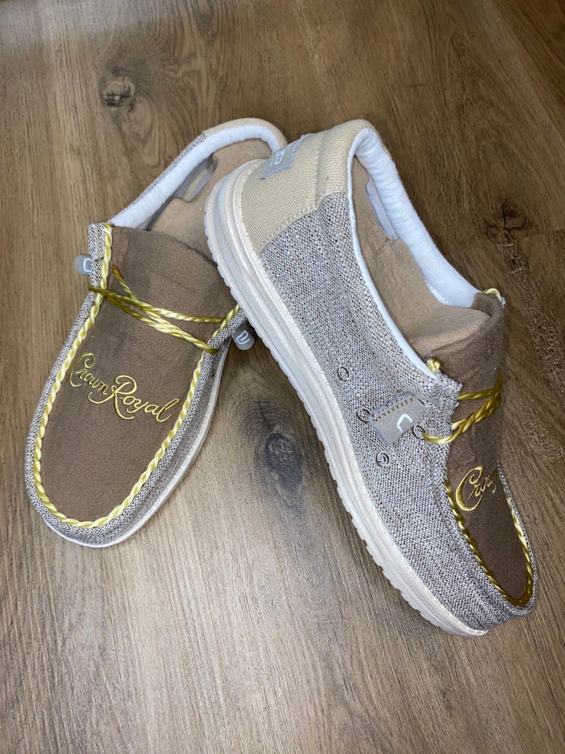 May include: A pair of brown and beige slip-on shoes with a gold embroidered logo that says "Crown Royal".