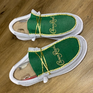 May include: A pair of white slip-on shoes with green fabric uppers and gold embroidery that reads "Crown Royal".