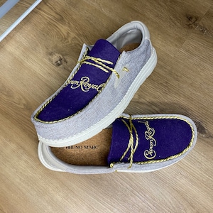 May include: A pair of gray canvas slip-on shoes with a purple suede upper and gold embroidery that reads "Crown Royal". The shoes have a white sole and a cork insole.