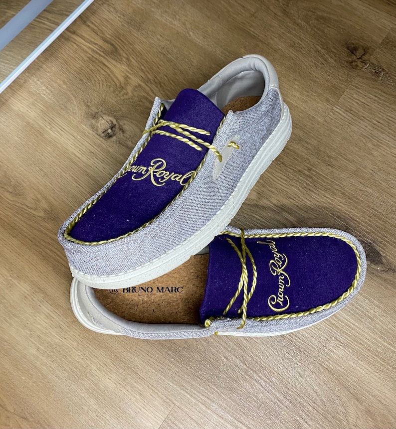 May include: A pair of gray canvas slip-on shoes with a purple suede upper and gold embroidery that reads "Crown Royal". The shoes have a white sole and a cork insole.