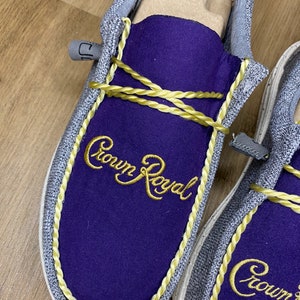 May include: A pair of gray canvas slip-on shoes with purple fabric lining and gold rope trim. The shoes feature the embroidered text "Crown Royal" in gold thread.