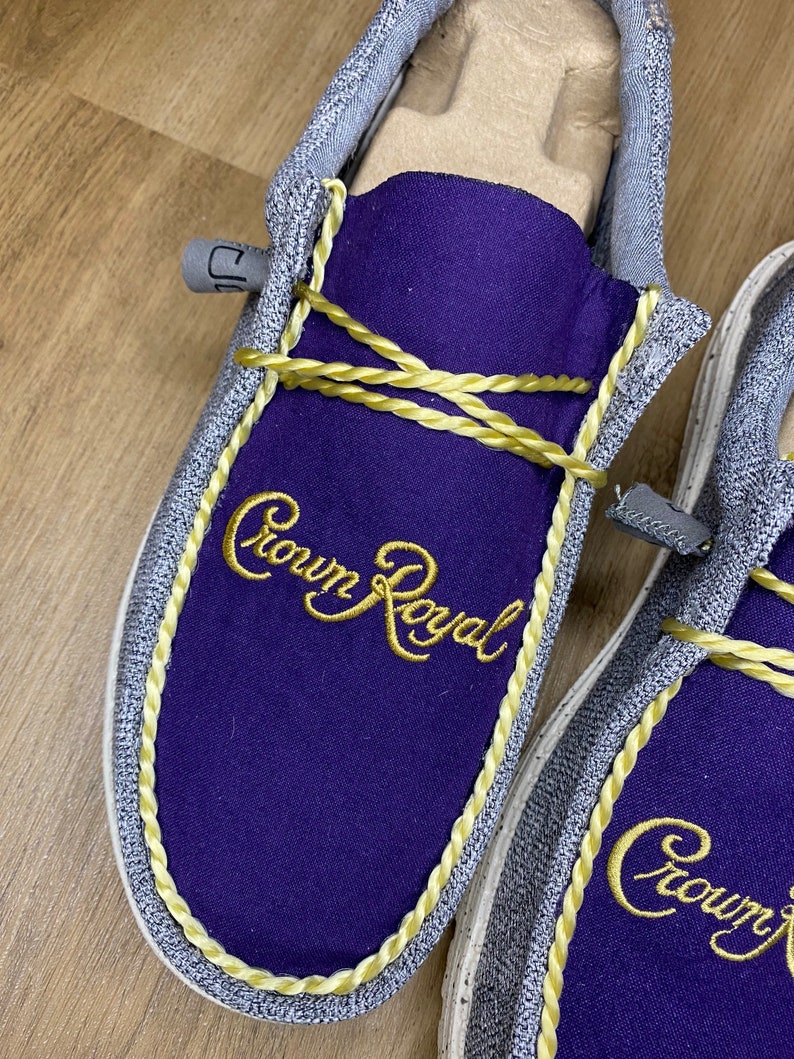 May include: A pair of gray canvas slip-on shoes with purple fabric lining and gold rope trim. The shoes feature the embroidered text "Crown Royal" in gold thread.