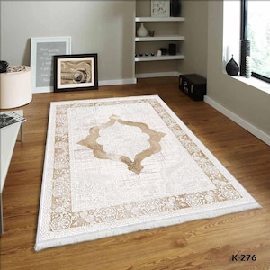 Pu&ograve; includere: Un tappeto beige e bianco con un grande disegno dorato intricato al centro. Il tappeto ha un bordo con frange.