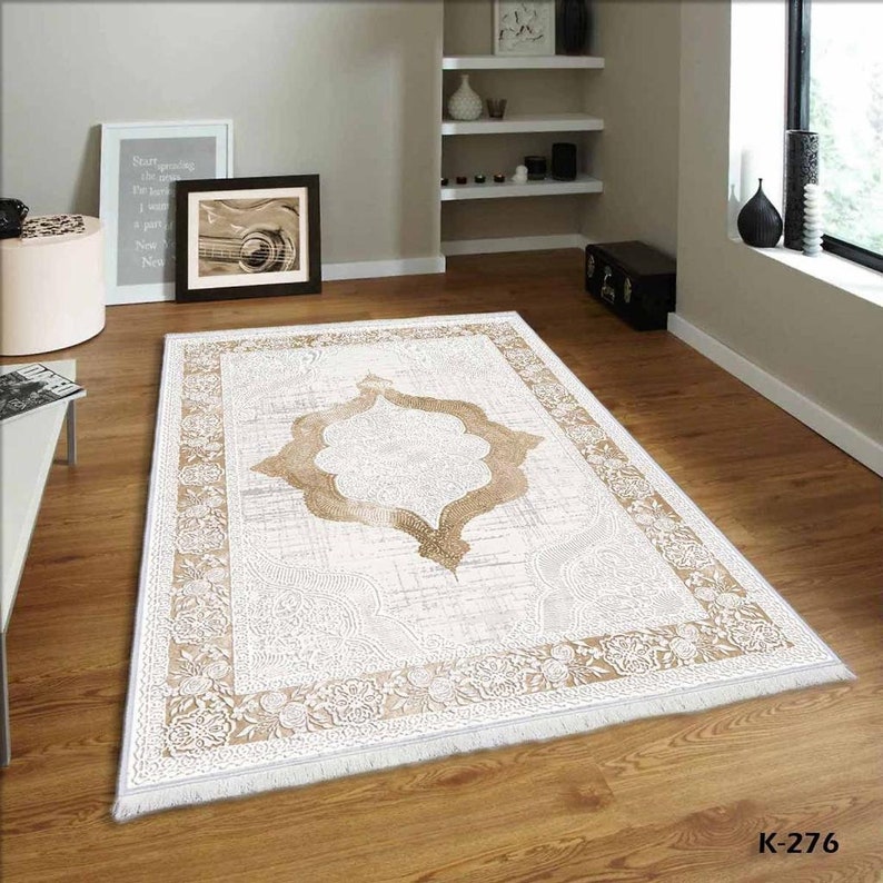 Pu&ograve; includere: Un tappeto beige e bianco con un grande disegno dorato intricato al centro. Il tappeto ha un bordo con frange.