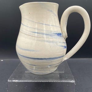 Puede incluir: Jarra de cerámica hecha a mano con base blanca y diseño de remolino azul. La jarra tiene un asa curva y un pico para verter. La cerámica se exhibe sobre un soporte acrílico transparente.