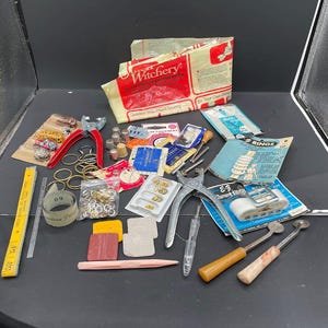May include: A collection of vintage sewing and jewelry making supplies, including a red pliers, a ruler, a spool of thread, a thimble, a package of rings, and a variety of beads and findings.