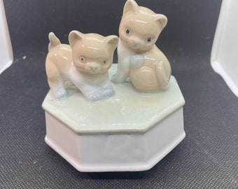 May include: A white porcelain music box with two beige kittens on top. The kittens are facing each other and the one on the right is holding a ball. The music box has a hexagonal base.