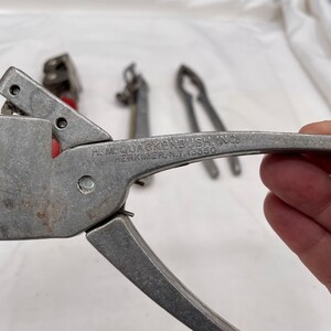 May include: A silver metal can opener with a black handle. The can opener is labeled "H.L. QUACKENBUSH INC. HERKIMER, N.Y. 13350".