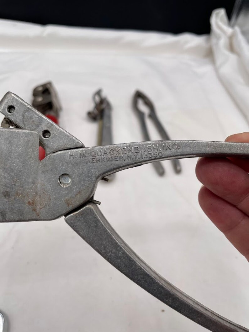 May include: A silver metal can opener with a black handle. The can opener is labeled "H.L. QUACKENBUSH INC. HERKIMER, N.Y. 13350".