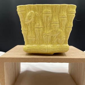 May include: A yellow-green ceramic planter with a textured bamboo design. The square-shaped planter has a speckled finish and sits on a wooden stand. The planter is suitable for small plants and adds a touch of nature to any space.