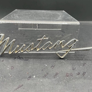 May include: A chrome "Mustang" emblem in a cursive font. The emblem has a stylized "flying horse" design at the end. The emblem is on a dark surface, with a clear plastic display case in the background.