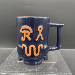 May include: A navy blue ceramic mug with a handle and a decorative base. The mug features orange abstract designs on the front. The mug is sitting on a clear surface.