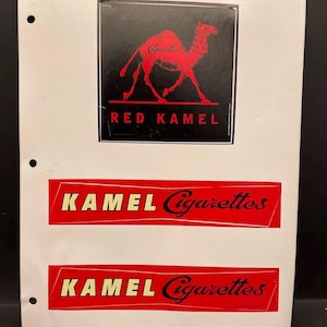 May include: A white sheet with three printed sections. The top section features a red camel silhouette on a black background with the text "RED KAMEL" below. Two red rectangles with "KAMEL Cigarettes" in white and black text are below.