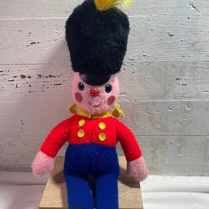 May include: A plush toy soldier wearing a black hat with a yellow plume, a red jacket with yellow buttons, blue pants, and black shoes. The toy is sitting on a wooden stool.